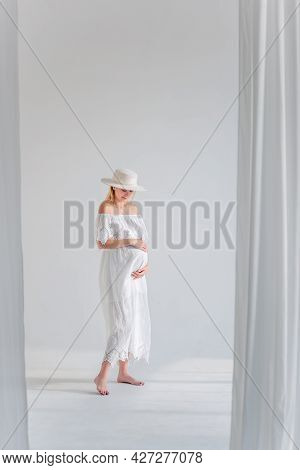 Young Pregnant Woman In Anticipation Of A Baby In A White Hat, Dress, Hugs Her Belly. Tender Blonde