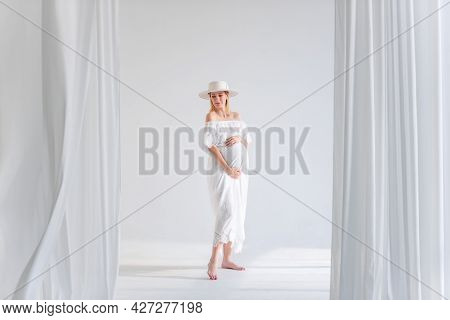 Young Pregnant Woman In Anticipation Of A Baby In A White Hat, Dress, Hugs Her Belly. Tender Blonde