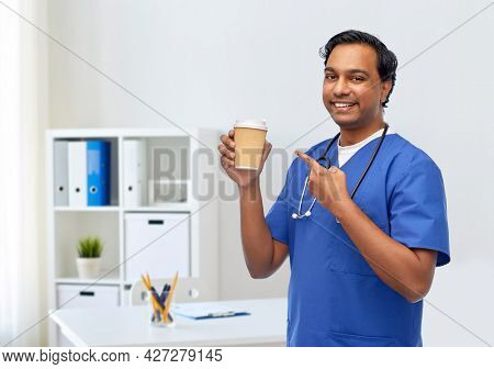 healthcare, profession and medicine concept - happy smiling indian doctor or male nurse in blue uniform with takeaway coffee cup and stethoscope over medical office at hospital background