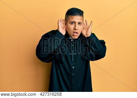 Young latin priest man standing over yellow background trying to hear both hands on ear gesture, curious for gossip. hearing problem, deaf 