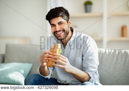 Cheat Meal Day. Happy Arab Man Eating Tasty Sandwich And Smiling To Camera, Sitting On Sofa In Livin