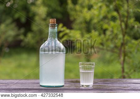 Big Bottle And Glass Of Moonshine Or Vodka On Wooden Table In Sunny Summer Day In Garden, Close Up. 