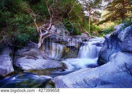 River Waterfall Between Rocks At Sunrise And Wooden Bridge In The Background. Navacerrada.