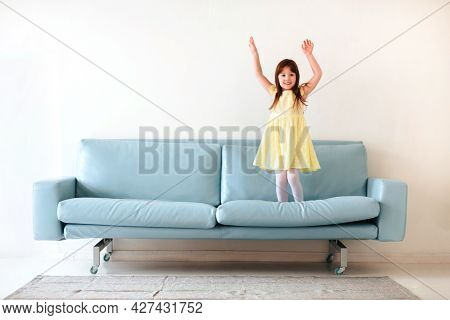 Little Caucasian Cute Lovely Girl In Yellow Dress Playing And Jumping On Blue Sofa In Living Room At