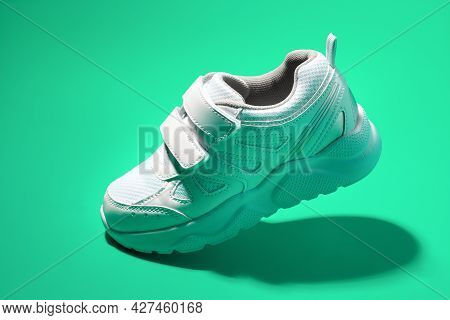 Side View Flying White Kid Sneaker With Velcro Fasteners Isolated On A Green Background With A Hard 