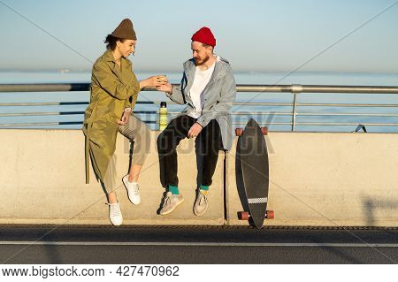 Happy Couple Drinking Hot Tea Outdoors Sitting On Bridge Broadwalk Dressed In Trendy Street Fashion 