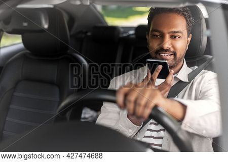 transport, people and technology concept - smiling indian man or driver driving car and recording message using voice command recorder on smartphone