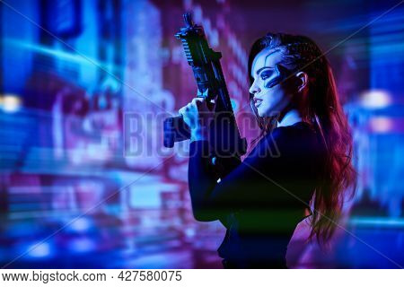 An attractive girl cyber warrior of the future stands on alert with an automatic gun among the neon lights of a night city. World of the future. Game, virtual reality.