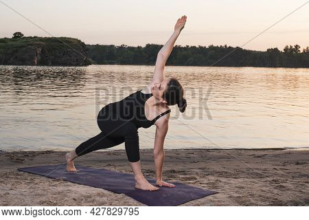 Girl Doing Yoga On A Mat On The Beach By The River At Sunset, Twisting In A Standing Position. Exten
