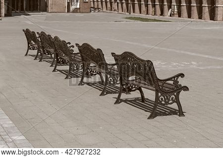 Row Of Empty Vintage Wooden Benches With Ornate Black Iron Forged Elements In Small Provincial Town 