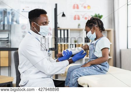 Adorable Little Afro Child Girl In Mask Sitting On The Couch In Medical Office And Looking At Her Ar