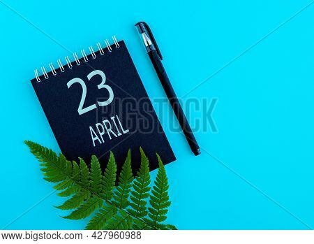 April 23th. Day 23 Of Month, Calendar Date. Black Notepad Sheet, Pen, Fern Twig, On A Blue Backgroun