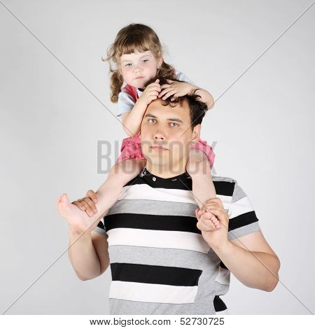 Little Girl Sits On Shoulders Of Her Father And Looks Down On Grey Background.