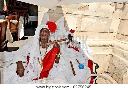 A smiling woman sitting on a bench in the street in a white dress with a huge cigar