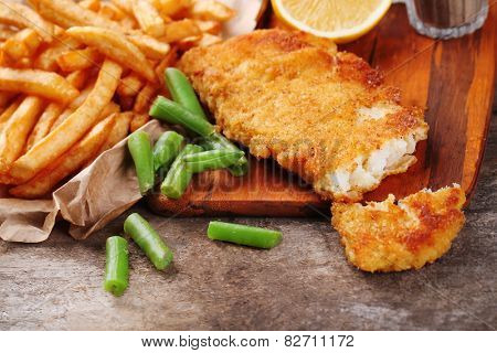 Breaded fried fish fillet and potatoes with asparagus and lemon on cutting board and rustic wooden background