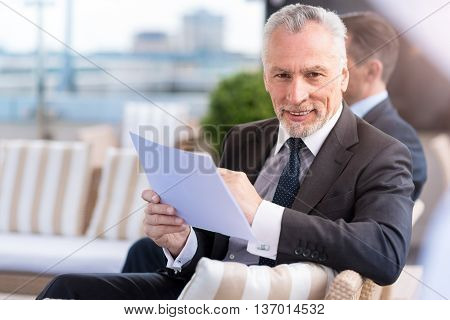 Work diligently. Positive senior, confident smiling man sitting on the couch and holding papers while his colleague working in the background