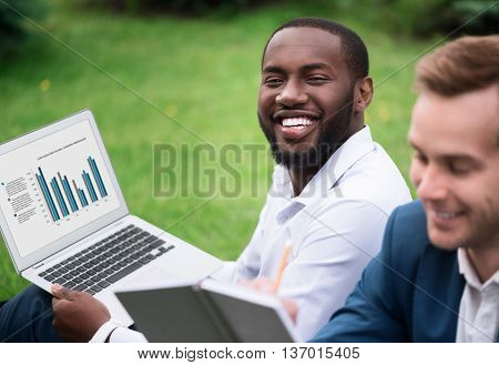 Evince your emotions. Cheerful delighted handsome smiling man sitting on the grass and holding laptop while resting with his colleague