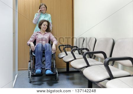 Nurse talking on phone and smile in hospital corridor, standing next to patient in wheelchair, holding handles