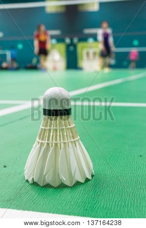 White badminton shuttlecock on a green floor with blurred players in badminton court.