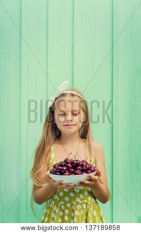 Beautiful blond girl on a background of turquoise wall holding a plate with cherry. Space for text