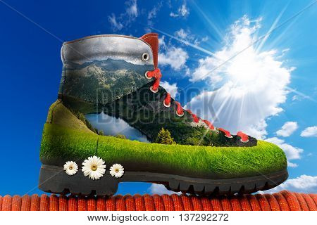 3D illustration of a hiking boot with mountain panorama and a lake, green meadow and three daisy flowers inside. On a blue sky with clouds and sun rays