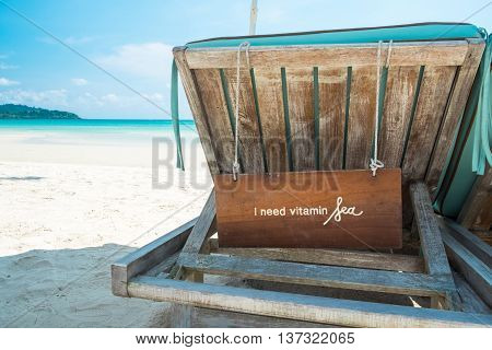 A wooden label with text " I need vitamin Sea " hanging behind the beach bed on a sunny day at the sea beach