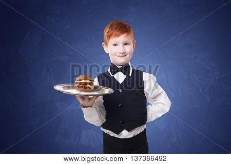 Little waiter stands with tray serving hamburger. Smiling redhead child boy in suit plays restaurant servant, gives burger at blue background