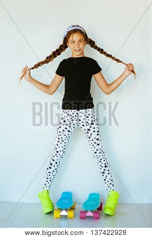 Pre teen girl wearing cool fashion clothing and sneakers posing with colorful skateboard against white wall