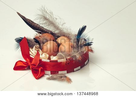 Many brown fresh eggs with black feathers and excelsior in round decorative box decorated by beautiful bright red bow on white background closeup studio