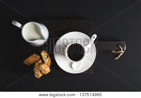 Cup of hot espresso, creamer with milk and cookies on dark rustic wooden board over black background, top view, horizontal composition