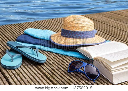 summer vacation blue water and accessories for beach holidays as straw hat, flip flops, turquoise towels and a book on a wooden bathing pier at the pool selected focus and narrow depth of field