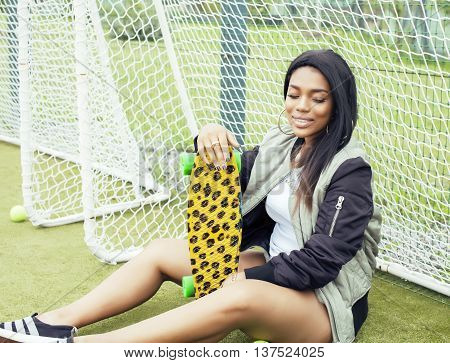 cool pretty smiling afro-american happy young woman with skate board hanging aroung at foothball field, lifestyle people concept