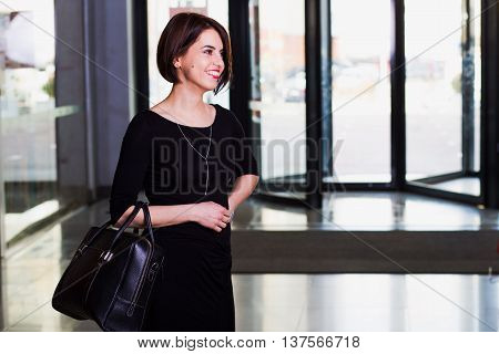 Office worker beautiful young businesswoman in black dress walking with briefcase in contemporary building. looking and smiling
