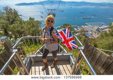 Happy tourist woman holding an English flag. On background the cable car that leads to Gibraltar Rock. Gibraltar is a British colony that is located at southern end of Iberian Peninsula.
