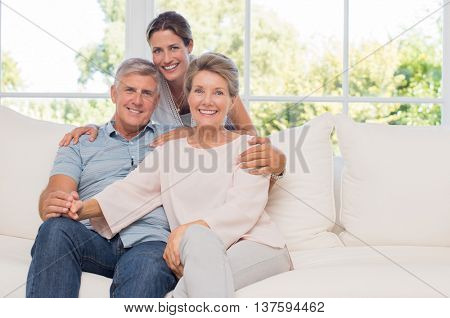 Portrait of senior couple sitting on couch with daughter. Elderly couple with daughter looking at camera. Mature daughter embracing her senior parents from behind.