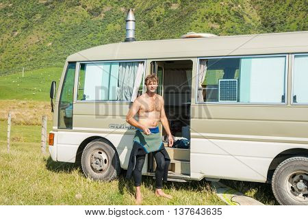 Tora,New Zealand -November 27,2011; Traveling alone in mobile home surfer stands by door to his converted bus with wetsuit pulled down to waist after morning surf.
