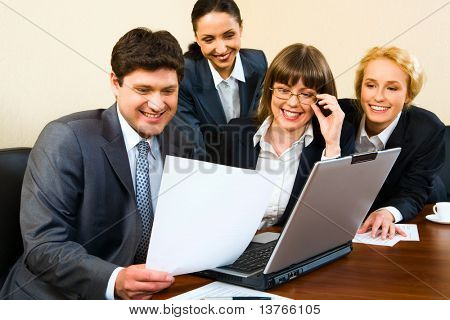 Group of four smiling businesspeople looking at the document in businessmanâs hand gathered together around the table with the laptop, white cup and papers on it