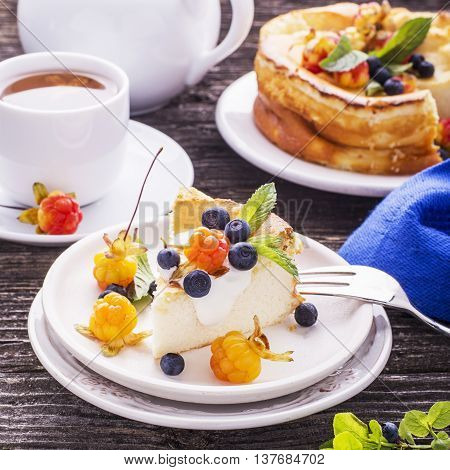 Homemade breakfast. Fresh cheesecake with Nordic berries cloudberries and blueberries in plain wooden background served with cloudberry tea in a white ceramic bowl. Decorated branches forest blueberries