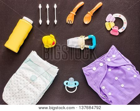Baby set of cloth diaper, disposable diaper, baby powder, tither, cotton buds, spoons, soother, nibbler and rubber duckling on dark background. Top view or flat lay