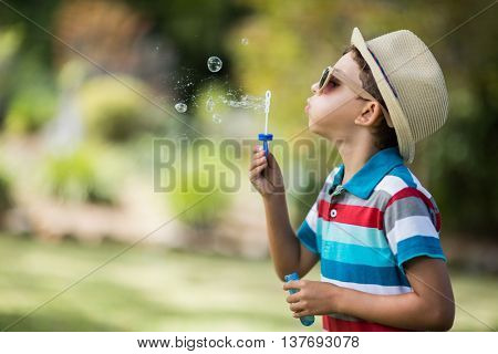Cute young boy in sunglasses blowing bubbles through bubble wand in park