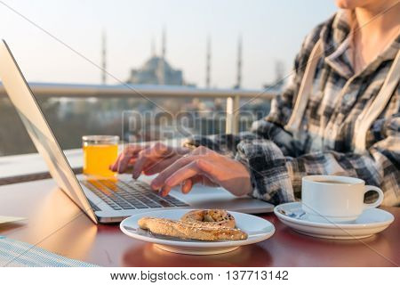 Working Breakfast Person Working on Laptop Computer at Cafe Terrace on Table with Coffee Mug Orange Juice and Bakery Cake Asian Urban Landscape on Background