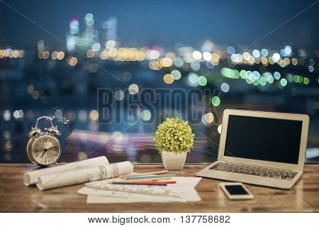 Front view of creative designer desktop with blank laptop stationery items plant alarm clock and smartphone on blurry night city background. Mock up