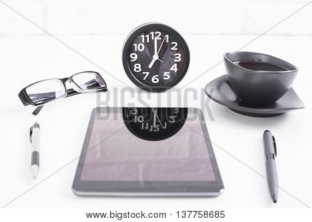 Closeup of white desktop with clock reflected on blank tablet screen pens glasses and coffee cup on white birck wall background. Mock up