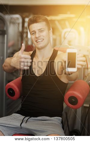 sport, bodybuilding, lifestyle, technology and people concept - smiling young man showing smartphone and thumbs up gesture in gym
