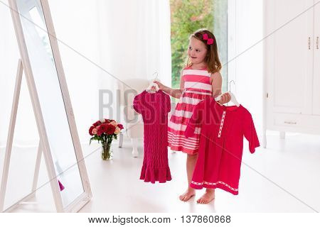 Little girl choosing dresses in white bedroom. Child watching mirror reflection holding pink dress choosing outfit. Girls nursery. Shopping clothing for kids. Dressing room interior for children.