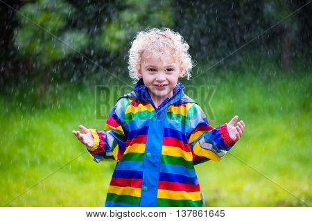 Little blond curly boy in a waterproof jacket in rainbow color playing in the rain. Kids having fun outdoors in autumn shower.