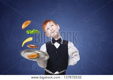 Little waiter dreaming with tray, serving hamburger. Cheeseburger levitates with separated toppings. Flying burger layers. Smiling redhead child boy in suit play restaurant servant at blue background