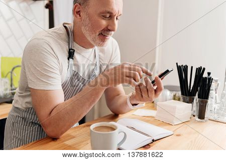 Time to relax. Cropped image of merry and positive male barista using smart phone while drinking coffee and making some notes being at work during his break