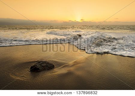 Beach sunset seascape is w detailed wave making it's way to the sandy shore as the sun sets with rays of light glowing on the horizon.