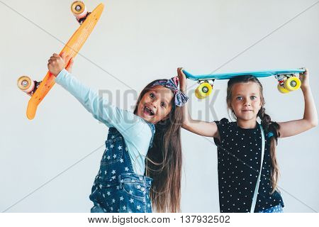 Two 7 years old children wearing cool fashion jeans clothing posing with colorful skateboard against white wall, urban style